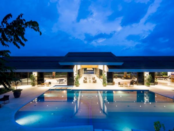 A luxurious open-air building with a large swimming pool, illuminated under a blue evening sky, surrounded by cozy seating areas.