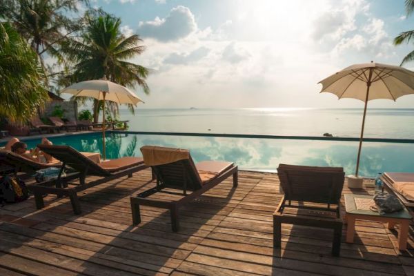 Poolside deck with lounge chairs, umbrellas, palm trees, overlooking a serene ocean view under a partly cloudy sky.