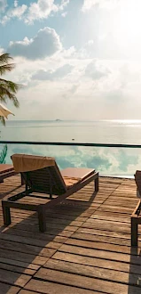 A tranquil poolside deck with lounge chairs, umbrellas, and palm trees overlooks a serene ocean under a cloudy sky.