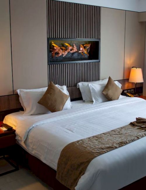 A cozy hotel room features a large bed with decorative pillows, warm lighting, and a modern wall fireplace.