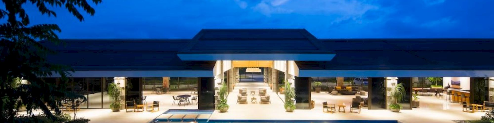 A modern building with an illuminated swimming pool at dusk, surrounded by seating areas and trees under a blue sky.