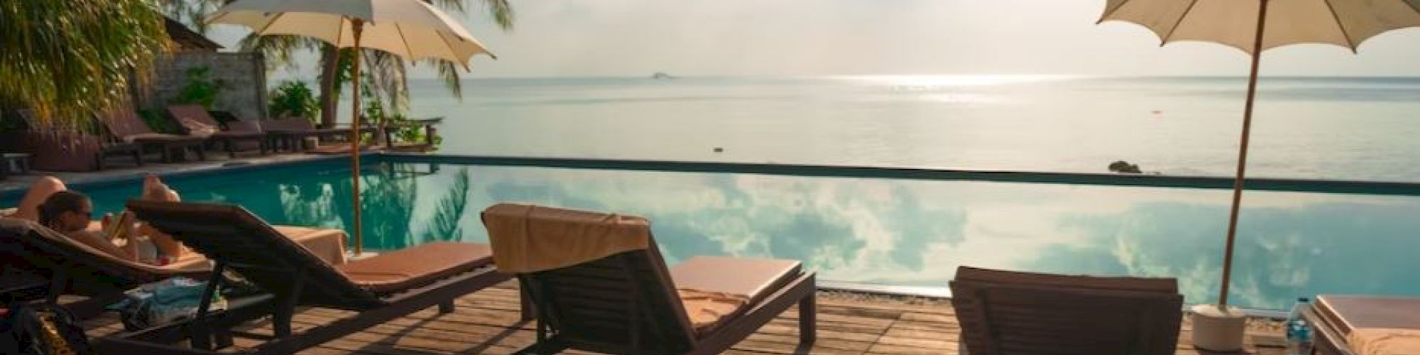 A serene poolside scene with lounge chairs, palm trees, and umbrellas under a bright sky by the water's edge.