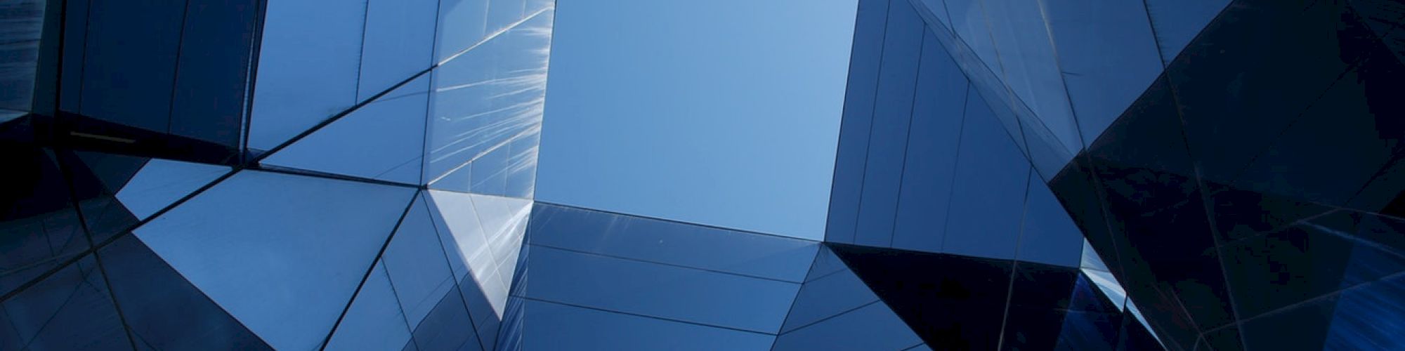 The image shows a modern architectural structure with reflective glass panels, forming a geometric pattern opening to the sky.