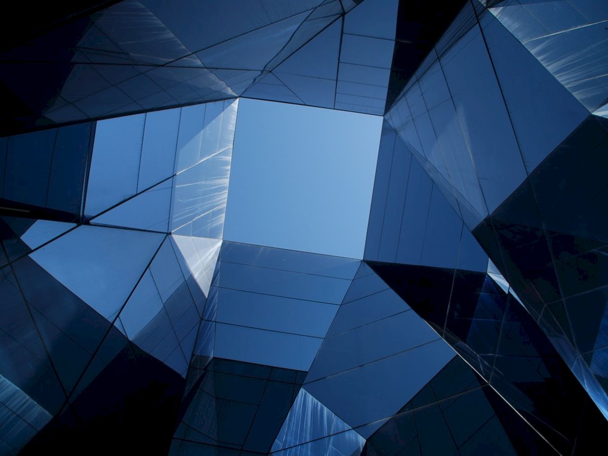 Geometric glass buildings viewed from below, forming a symmetrical pattern with the sky visible through the center.