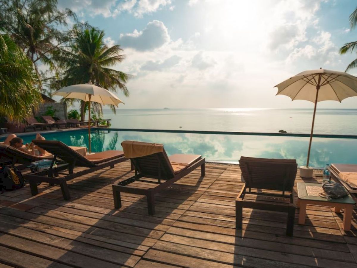 A serene poolside view with lounge chairs, umbrellas, palm trees, and a distant ocean horizon under a partly cloudy sky.
