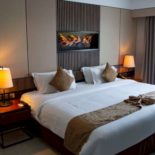 A cozy hotel room features a large bed with pillows, bedside lamps, decorative wall art, and a window with drawn curtains.