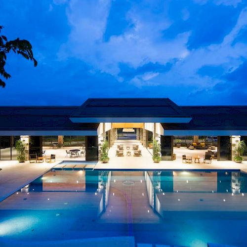 A luxurious, illuminated pool area at dusk with a modern, open-design building in the background, surrounded by tropical plants.