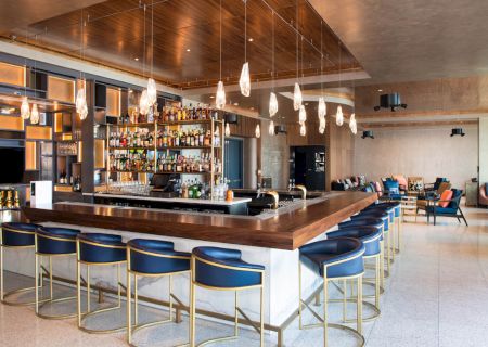 A stylish bar with a central counter, surrounded by blue bar stools, featuring shelves stocked with bottles, and modern overhead lighting.