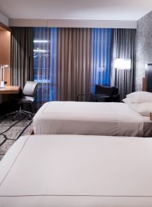 The image shows a modern hotel room with two beds, large TV, desk, and stylish decor.