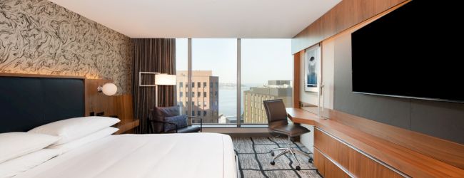 A modern hotel room with a large bed, desk, chair, TV, and a window view of city buildings and water.