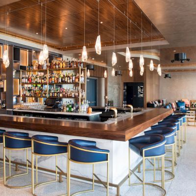 A modern bar with blue bar stools, hanging lights, and a stocked shelf of bottles, alongside an open seating area with couches and chairs.