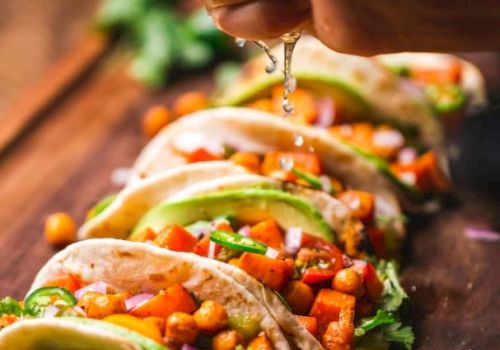 The image shows a hand squeezing lime juice over a row of tacos filled with chickpeas, vegetables, and garnished with herbs on a wooden board.