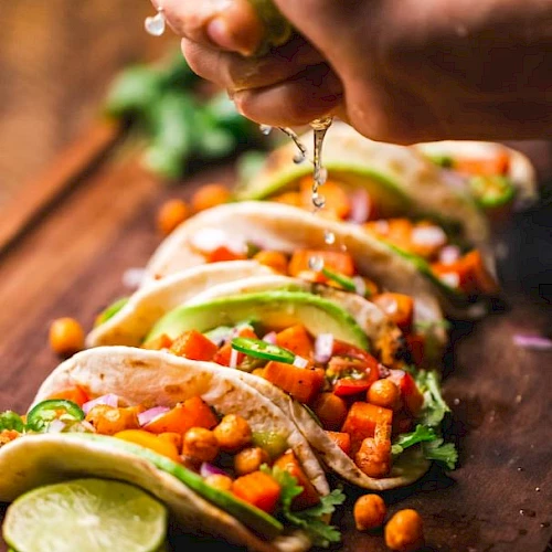 The image shows a hand squeezing lime juice over a row of tacos filled with chickpeas, vegetables, and garnished with herbs on a wooden board.