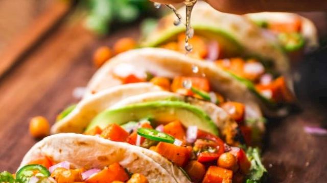 The image shows a hand squeezing lime juice over a row of tacos filled with chickpeas, vegetables, and garnished with herbs on a wooden board.