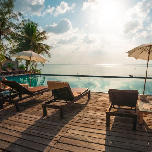 Sun loungers and umbrellas by a pool facing the ocean, with palm trees and a sunny sky in the background.