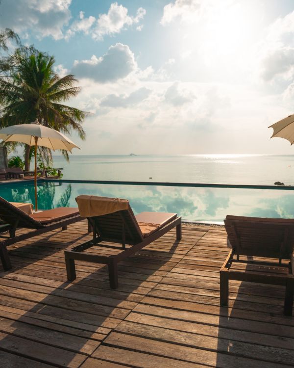 Sun loungers and umbrellas by a pool facing the ocean, with palm trees and a sunny sky in the background.