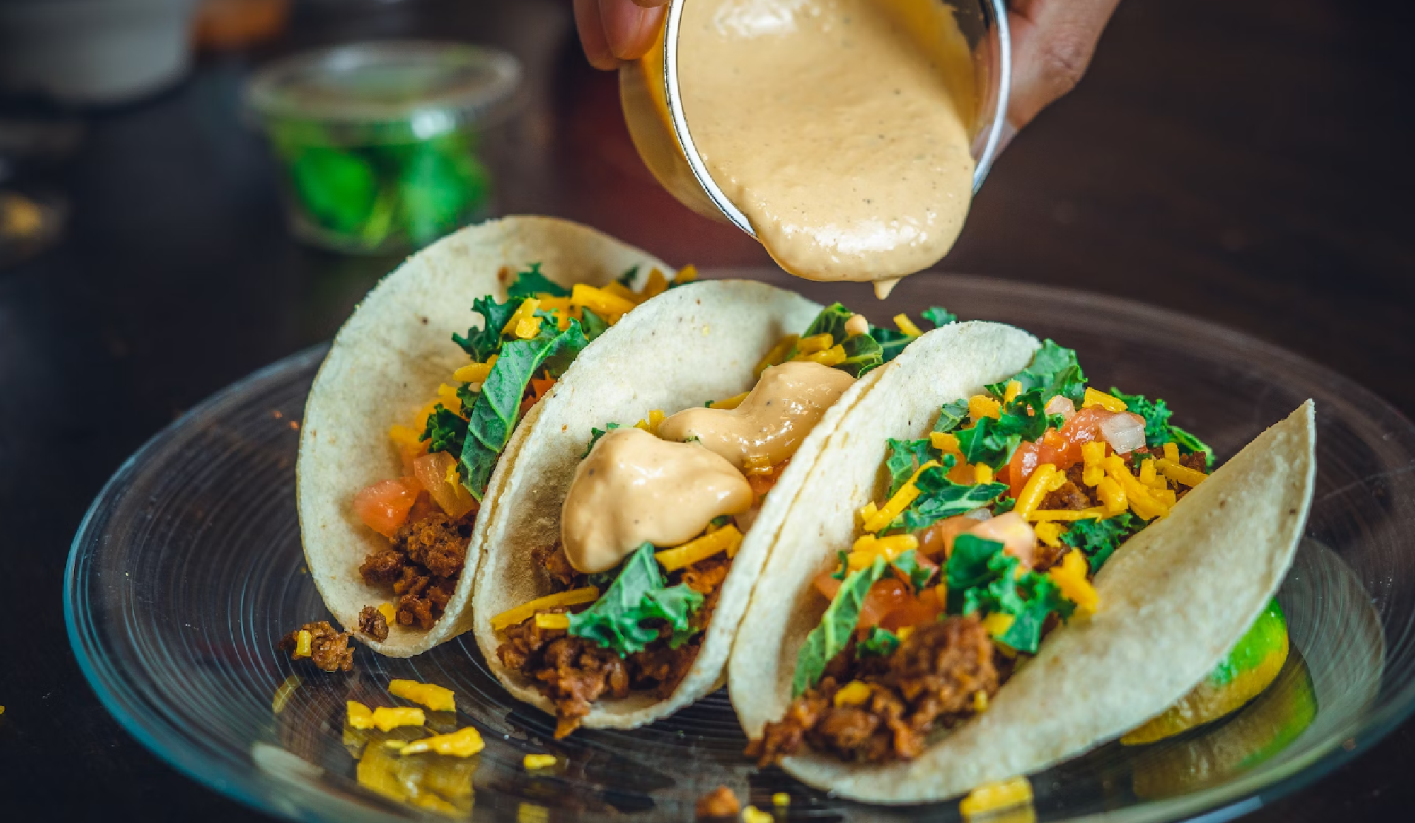 Three tacos with beef, cheese, kale, and tomatoes are being topped with sauce on a glass plate.