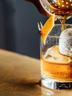 A hand is pouring a drink through a strainer into a glass with ice and a lemon twist garnish, on a wooden surface.