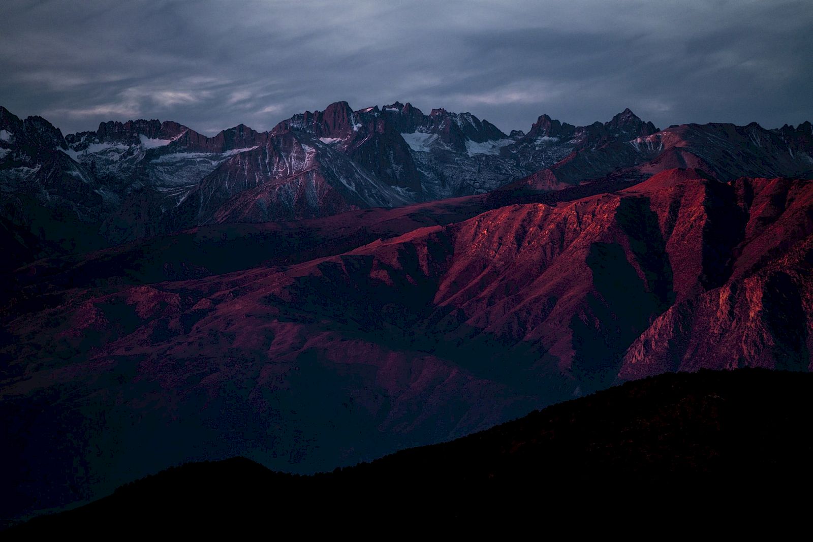 A mountain range is bathed in a dramatic sunset glow, with jagged peaks and deep shadows enhancing the scene's beauty and mood.