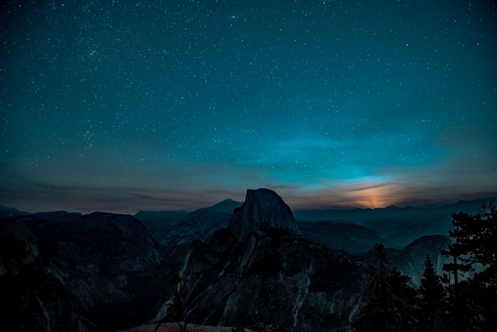 A starry night sky over a silhouetted mountain landscape with a faint glow, possibly from the moon or sunset, on the horizon.