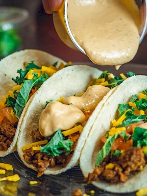 Three tacos filled with ground meat, lettuce, cheese, and tomatoes are being topped with a creamy sauce.