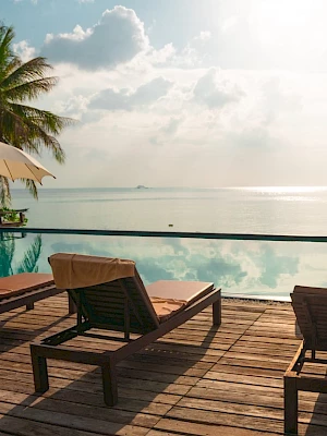 A tranquil seaside scene with lounge chairs, umbrellas, a pool, and palm trees overlooking the ocean under a partly cloudy sky.