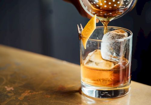A drink is being poured into a glass with ice and a lemon twist garnish on a counter.