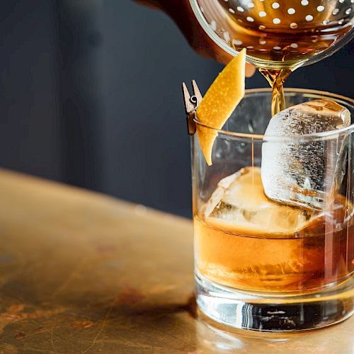 A drink is being poured into a glass with ice and a lemon twist garnish on a counter.
