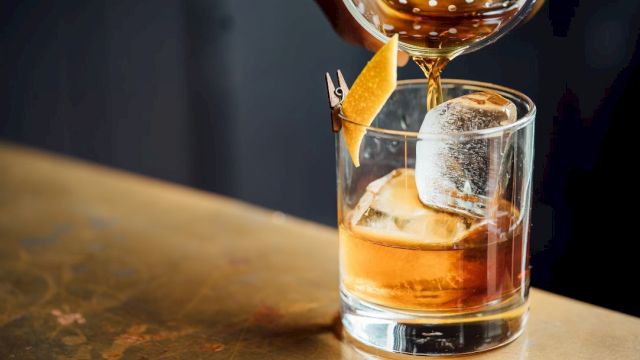 A drink is being poured into a glass with ice and a lemon twist garnish on a counter.