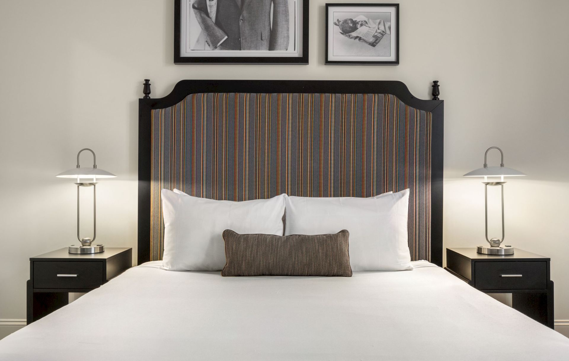 A neatly made bed with white pillows and a striped headboard, flanked by lamps on bedside tables, with framed photos above.