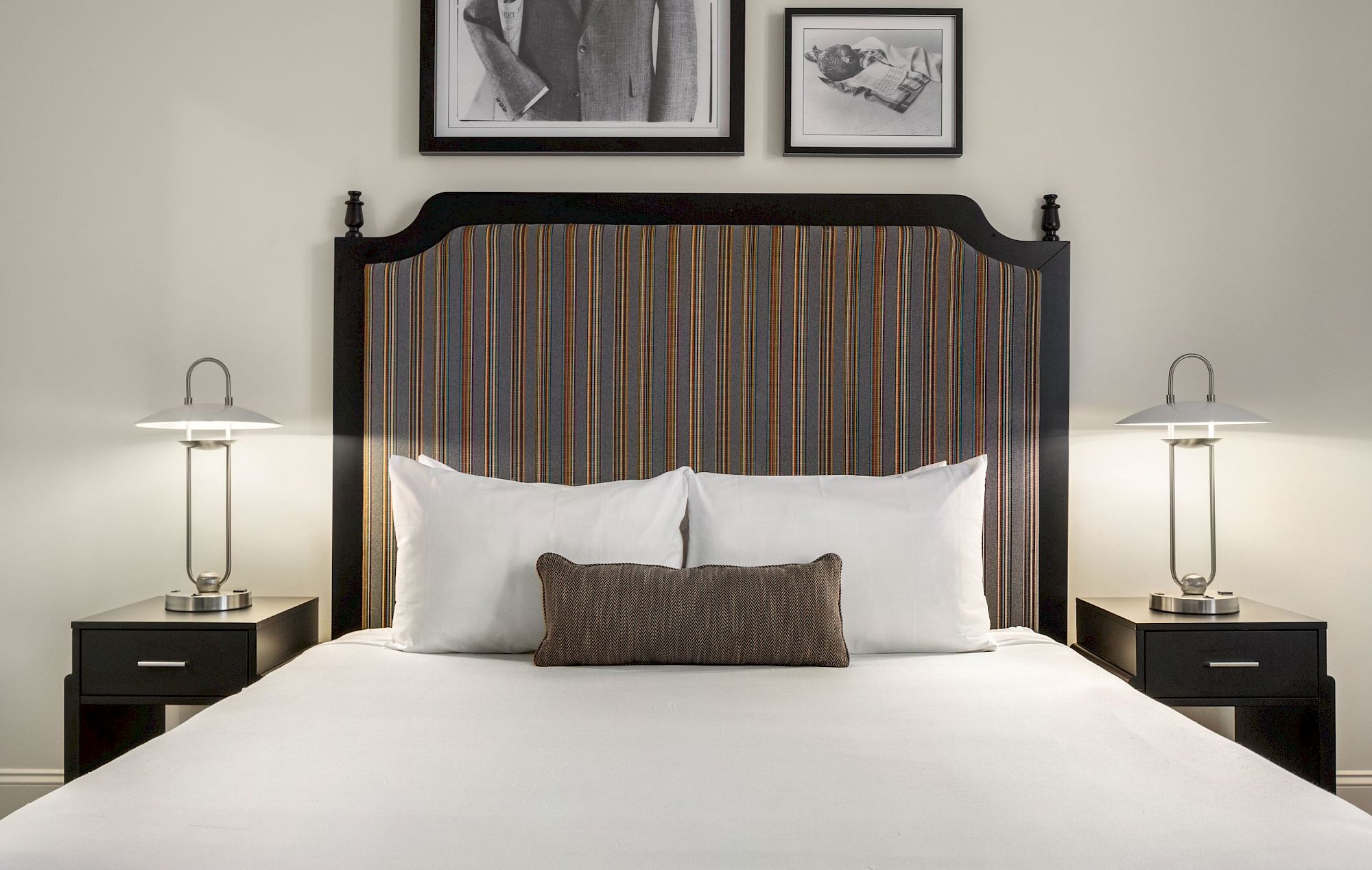 A neatly made bed with white pillows and a striped headboard, flanked by lamps on bedside tables, with framed photos above.