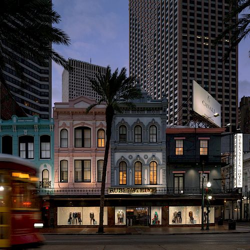 A streetcar passes colorful historic buildings at dusk, with palm trees and a tall skyscraper in the background.