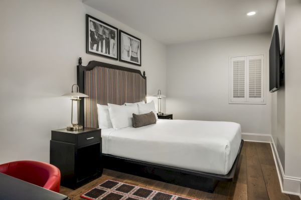 A modern hotel room with a double bed, nightstands with lamps, framed art, a wall-mounted TV, and a small desk with a red chair.