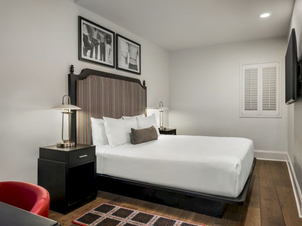A modern hotel room with a double bed, nightstands with lamps, framed art, a wall-mounted TV, and a small desk with a red chair.