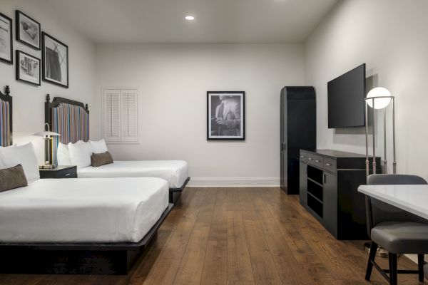 A modern hotel room with two beds, wall art, a flat-screen TV, desk, chair, and wood floor, creating a cozy and stylish ambiance.