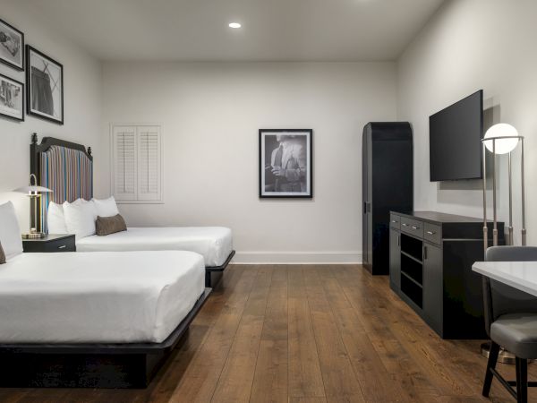 A modern hotel room with two beds, wall art, a flat-screen TV, desk, chair, and wood floor, creating a cozy and stylish ambiance.