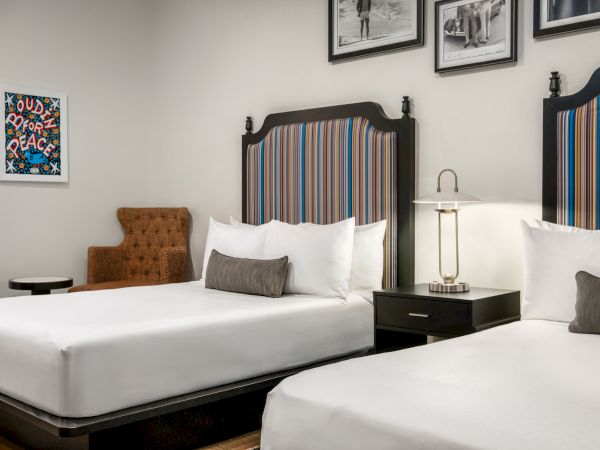 The image shows a hotel room with two beds, striped headboards, framed photos, a brown chair, a side table, and a lamp on a nightstand.