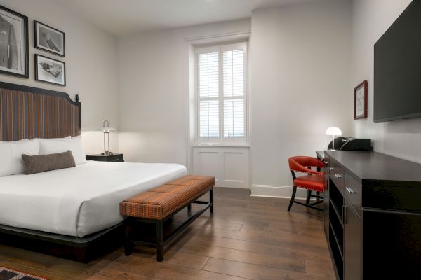 A modern hotel room with a bed, a bench, a desk with a lamp, and a flat-screen TV, featuring wooden flooring and framed art.