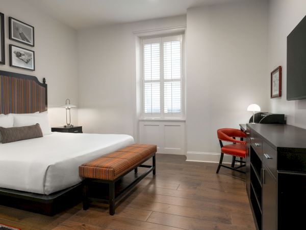 A modern hotel room with a bed, a bench, a desk with a lamp, and a flat-screen TV, featuring wooden flooring and framed art.