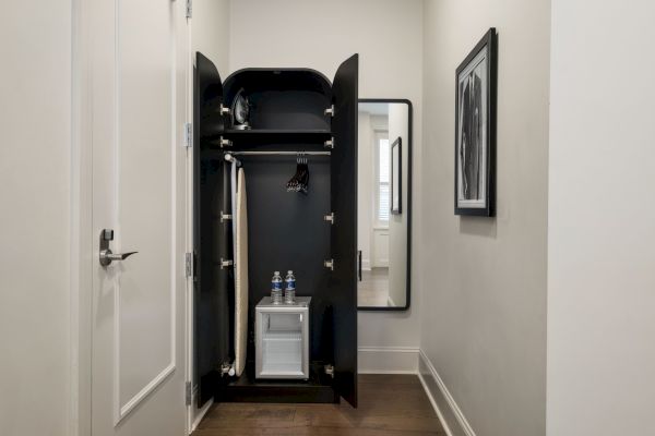 A closet with a built-in fridge is open, showing two water bottles and a hanging garment. A mirror and framed picture are on the wall.
