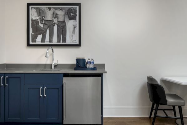 A small kitchenette features blue cabinets, a sink, mini-fridge, and two water bottles. A black-and-white photo hangs above. A chair is nearby.