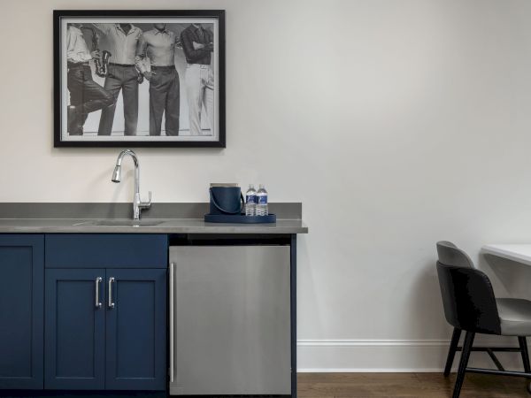 A small kitchenette features blue cabinets, a sink, mini-fridge, and two water bottles. A black-and-white photo hangs above. A chair is nearby.
