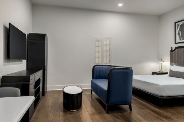 The image shows a modern room with a bed, blue couch, TV, and small round table on a wooden floor, under soft lighting.