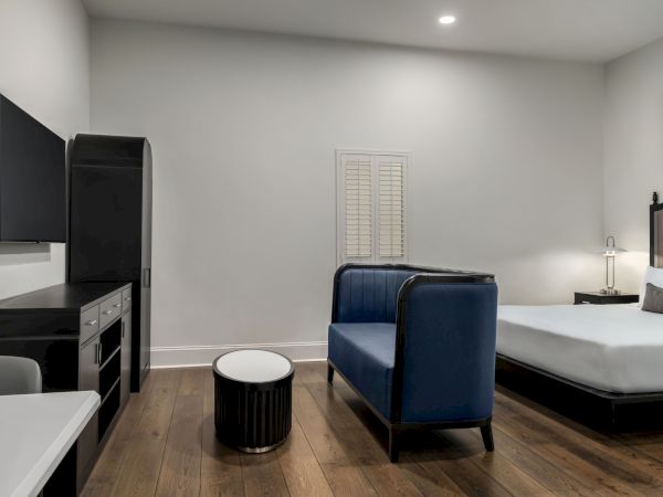 The image shows a modern room with a bed, blue couch, TV, and small round table on a wooden floor, under soft lighting.
