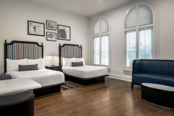 The image shows a hotel room with two beds, striped headboards, framed pictures, a small table, and a blue sofa near arched windows.