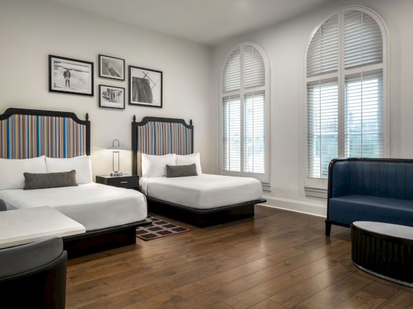 The image shows a hotel room with two beds, striped headboards, framed pictures, a small table, and a blue sofa near arched windows.