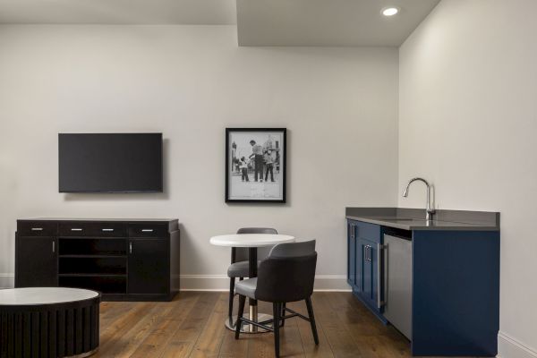The image shows a modern room with a TV, wall art, small table with chairs, cabinet, and sink, all set on a wooden floor.