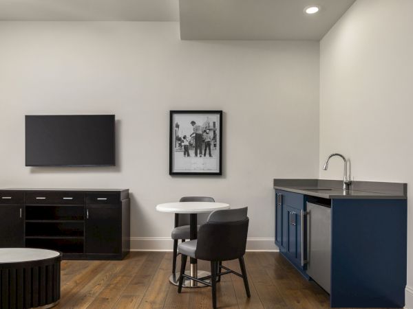 The image shows a modern room with a TV, wall art, small table with chairs, cabinet, and sink, all set on a wooden floor.
