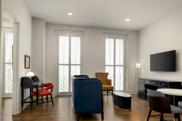 A modern living room with sofas, a TV, a desk with chairs, a coffee table, and large windows with blinds on hardwood flooring.