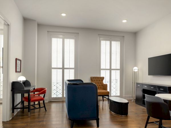 A modern living room with sofas, a TV, a desk with chairs, a coffee table, and large windows with blinds on hardwood flooring.